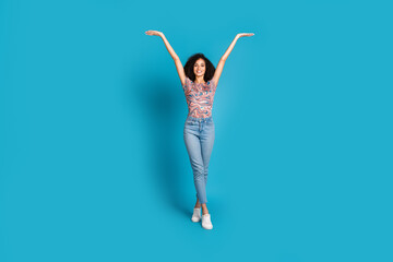 Joyful young woman with curly hair raising hands in casual style, expressing happiness against a...