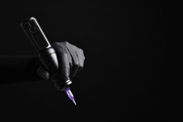 Professional artist in gloves holding tattoo machine on black background, closeup. Space for text