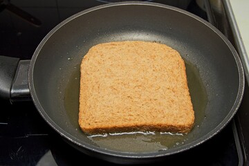 A slice of whole wheat bread is being fried in a pan with oil. The bread is partially submerged in the oil, which is bubbling around the edges.