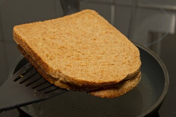a close-up of a sandwich being cooked in a frying pan. A spatula is lifting the sandwich, which consists of two slices of brown bread. This image is relevant for illustrating a cooking process,.