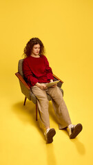 Boring young lady, with curly hair, wearing casual clothes, sitting in chair with tablet against yellow studio background. Concept of human emotions, self-expression, fashion and style.