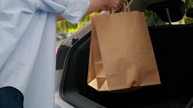 Unrecognizable Caucasian senior customer person lady elderly female shopper buyer put packages paper bags load purchases in car trunk automobile electric vehicle shopping delivery outside city street