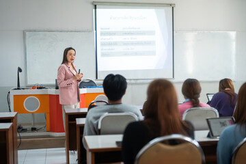 A lecturer stands at the front of the classroom, engaging students with a presentation projected on the screen. The students sit at desks, focused on the lecture.