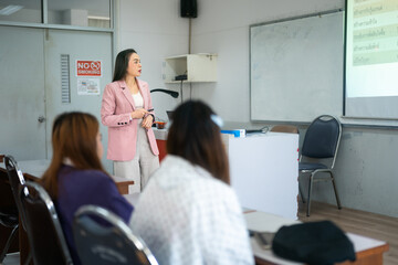 A lecturer stands at the front of the classroom, engaging students with a presentation projected on the screen. The students sit at desks, focused on the lecture.