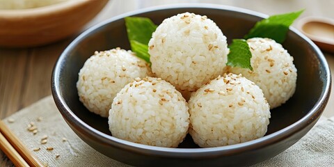 White sesame rice balls arranged in a dark bowl with green mint leaves on a light textile surface, evoking traditional Japanese cuisine and flavors.