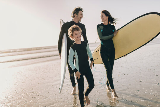 Family in wetsuits walking with surfboards on the beach