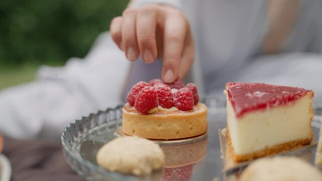 A womans hand gently picks a fresh raspberry from beautifully arranged dessert plate during picnic. Scene emphasizes warmth, simplicity, and enjoyment of treats in serene garden setting