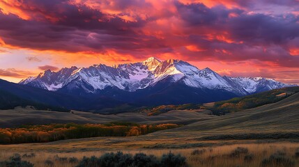 Obraz premium A vibrant sunset with a mountain range in the background, covered in snow. The sky is filled with dramatic clouds, and the landscape in the foreground features grasslands with a few trees.