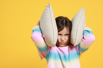 Annoyed little girl covering her ears with pillows from loud sound on yellow background. Space for...