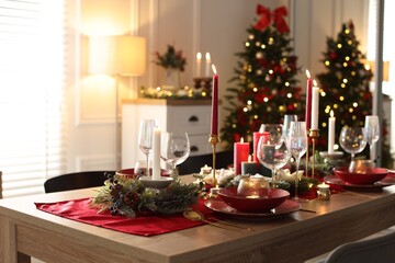 Christmas table setting with stylish dishware, glasses and burning candles in festive decorated room