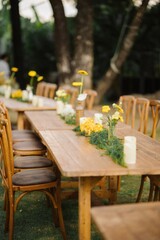 A rustic outdoor dining setup featuring wooden tables adorned with yellow flowers and greenery, creating a warm and inviting atmosphere.