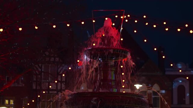 Fountain flowing brightly at night, illuminated by glowing red string lights. Water cascading down tiers, sparkling under festive evening lights. Ornamental fountain surrounded by decorations