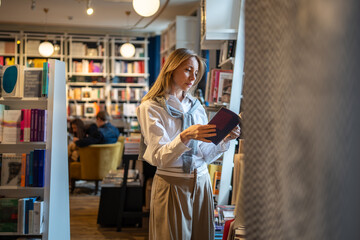 Thoughtful woman customer chooses and reads books from shelves in bookshop. Book worm female and reading hobby, browsing literature, book lover, looking for textbooks for study as adult student