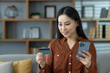 Asian woman using phone and card for online shopping at home. Relaxed on sofa, she engages in...