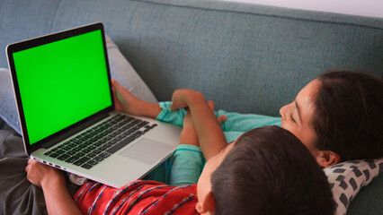 Fototapeta premium Rear View Shot Of Two Children Sitting On Sofa At Home Using Green Screen Laptop Computer Together