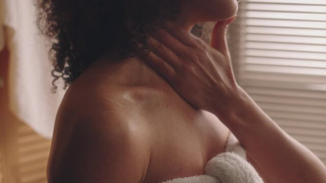 Side closeup shot of young curly-haired Biracial lady in towel rubbing moisturizing body lotion on shoulders and neck after shower, skincare and beauty routine