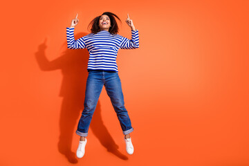 Full length photo of adorable lovely girl wear striped shirt jumping high pointing two fingers up empty space isolated orange color background