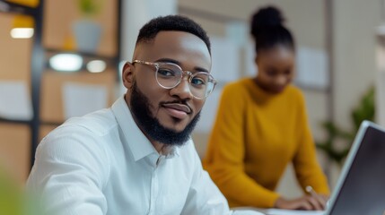 Obraz premium Young man with glasses and well-groomed beard works at desk in modern office while colleague collaborates in background