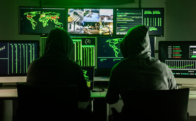 Two hackers working on computers showing world maps and security camera footage