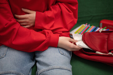 A teenage girl takes a sanitary pad out of her backpack, holds onto her stomach, which hurts