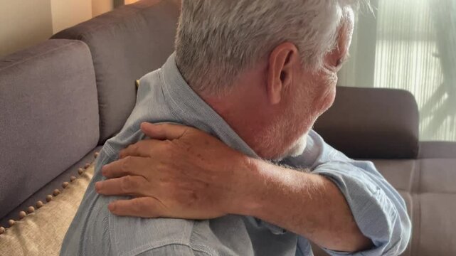 Mature retiree sitting on a couch, massaging his neck and shoulder to relieve pain caused by aging, stress, and physical discomfort, while also dealing with back pain, exhaustion, and other aging