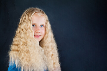 Cheerful blond young girl with long healthy curly hair on black background, fashion portrait