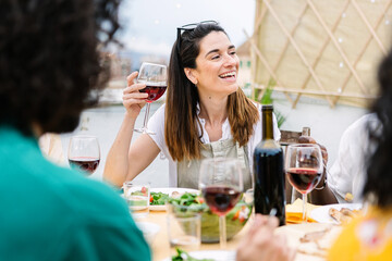 Happy group of multiracial young friends having dinner at rooftop summer party. Multiracial people laughing and having fun while drinking and eating sitting on table during celebration
