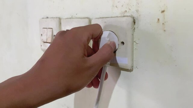Young adult man hand holding and unplug white electrical plug in wall outlet socket at home. Closeup. Side view.