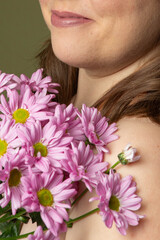 Fototapeta premium Curvy woman with clear skin imperfections in studio, purple daisy flowers and green background, unrecognizable