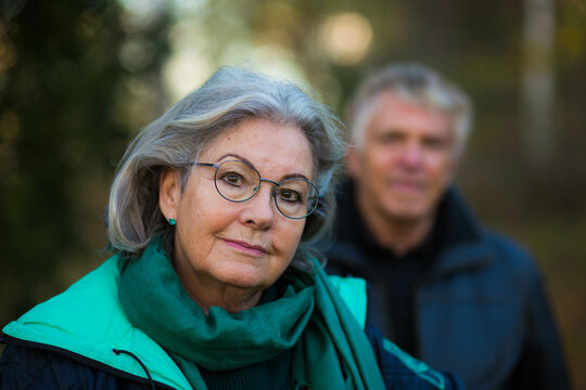 A portrait of an older woman and her husband, out of focus, in the background outside in the Swedish nature
