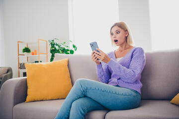 Young woman with blond hair on sofa holding smartphone looking surprised sitting in brightly lit modern living room