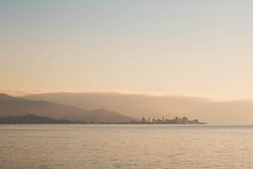 Fototapeta premium A distant view of Batumi city at sunset, with its modern skyline silhouetted against the hazy mountains and calm sea, under a warm golden sky. Concept of urban landscapes and travel