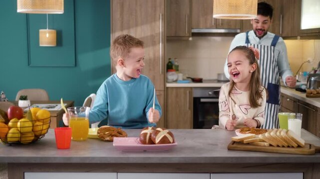 Happy children brother and sister shouting, having fun, and pounding fists with forks and knives on the table while waiting for their father to serve breakfast in the kitchen.