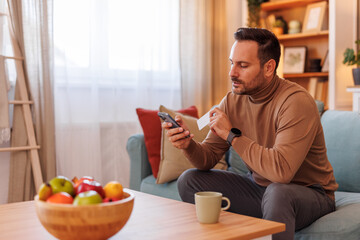 Serious businessman ordering online over mobile apps with credit card while sitting on sofa at home