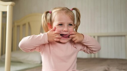 Little Girl joking sticking tongue out with silly expression. Child making face. Funny foolishness portrait of baby girl laughing in bedroom. Adorable Caucasian kid doing crazy comical gesture at home