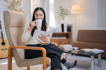 Young Asian woman sitting comfortably in an armchair, sipping coffee from a mug while engrossed in...
