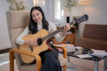 Happy young asian woman sitting on armchair and playing her acoustic guitar at home, with some books and musical headphones on the table next to her