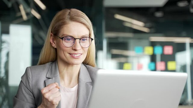 Satisfied happy businesswoman in jacket reading good news on laptop sitting at desk at workplace in business office. Smiling glad woman smiles, rejoices at a positive message on the computer. Close up