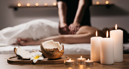 Close-up of a coconut with a tropical flower, candles, and incense in a modern spa. In the background, a young woman is receiving a body scrub massage. Relaxation and body care concept.