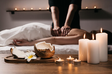 Close-up of a coconut with a tropical flower, candles, and incense in a modern spa. In the background, a body scrub massage is being performed on a young woman. Spa, relaxation, and body care concept.