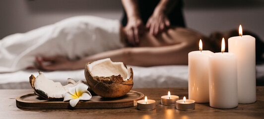Close-up of a coconut with a tropical flower, candles, and incense in a modern spa. In the background, a body scrub massage is being performed on a young woman. Spa, relaxation, and body care concept.