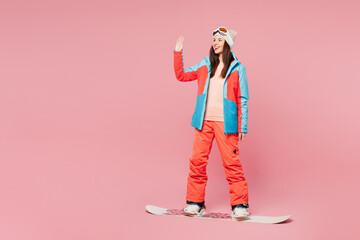 Full body young woman wear warm blue padded jacket ski goggles mask spend weekend winter season in mountains riding snowboard waving hand greet meet isolated on plain pink background. Hobby concept. © ViDi Studio