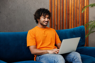 Young IT Indian man wear orange casual clothes sit on blue sofa couch hold use work on laptop pc...