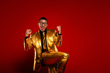 Charismatic young man poses joyfully in a shiny gold suit with a vibrant red background, embodying...