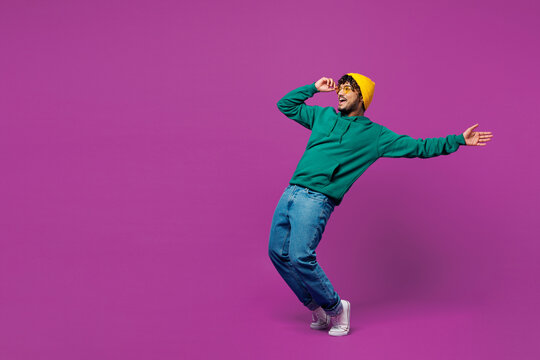 Full body side view young shocked fun Indian man in green hoody yellow hat sunglasses casual clothes lean back stand on toes look aside on area isolated on plain purple background. Lifestyle concept