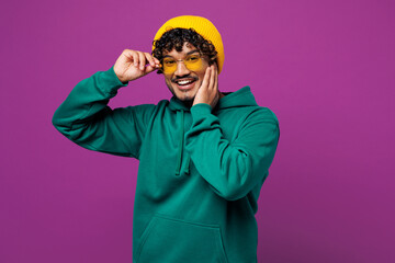 Young smiling cheerful satisfied happy Indian man he wearing green hoody yellow hat casual clothes touch sunglasses look camera isolated on plain purple background studio portrait. Lifestyle concept.