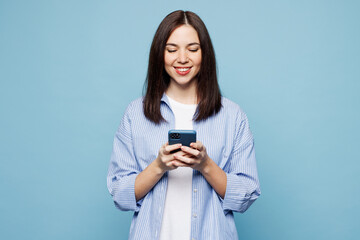 Young smiling Caucasian woman she wears shirt casual clothes hold in hand use mobile cell phone chatting online isolated on plain pastel light blue cyan background studio portrait. Lifestyle concept.