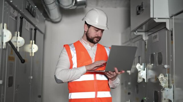Professional electrical engineer works inside server room. Bearded man types data on laptop, closes, gives thumbs up and looks at camera. Inside industrial hall or factory.