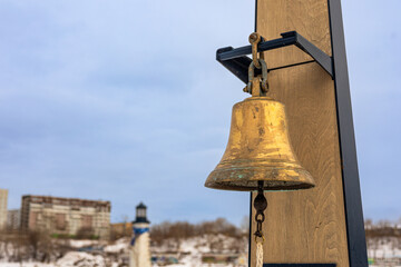 Obraz premium Brass bell hanging from a wooden frame. Ships bell, rynda. Defocused snowy landscape with lighthouse and buildings on cold full winter day.