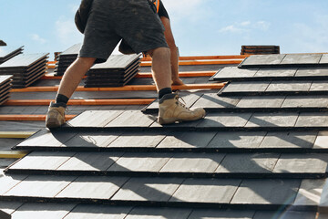 Roofing workers install slate tiles on a residential home under a clear blue sky during the day